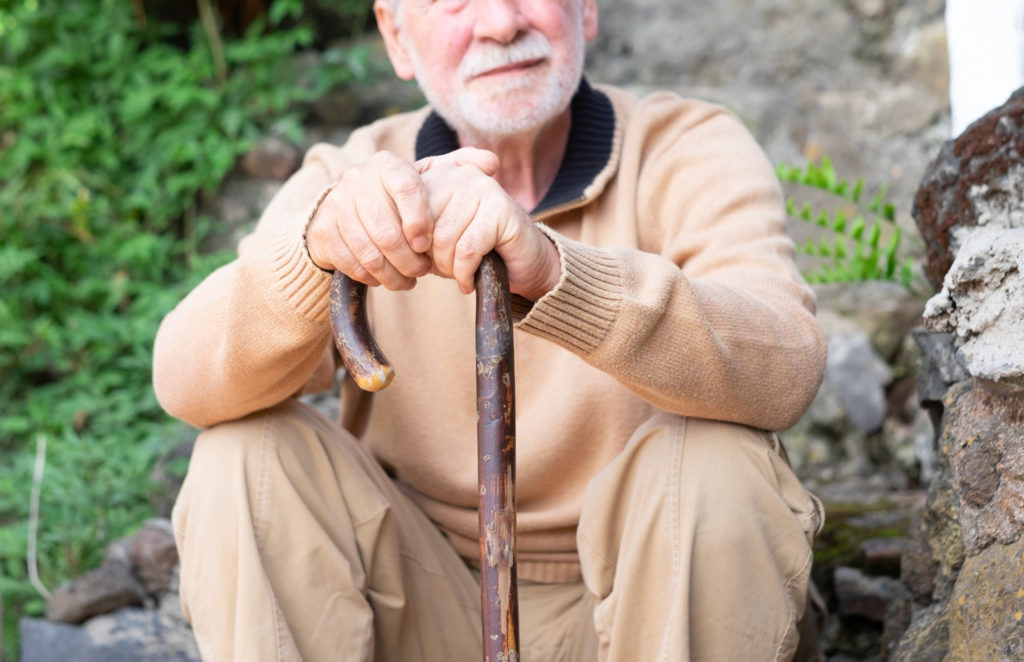 Un homme âgé est assis sur des marches à l'extérieur, et s'appuie sur une canne en bois de ses deux mains