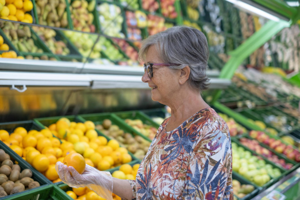 Une femme âgée tient une orange devant un rayon de fruits et légumes dans un supermarché