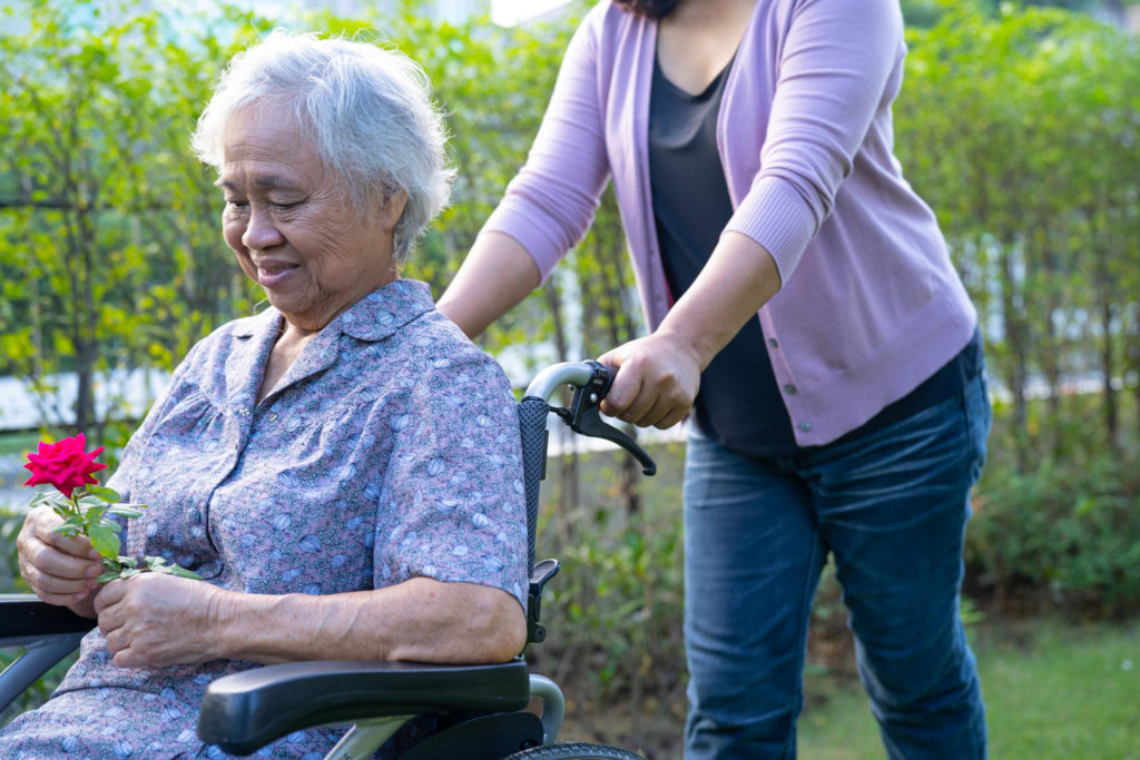 Une femme pousse une personne âgée tenant une rose avec un fauteuil roulant dans un parc