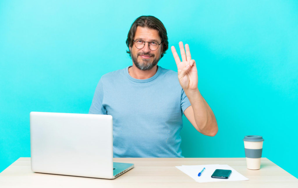Un homme assis à un bureau avec un ordinateur devant lui fait trois avec les doigts sur un fond bleu